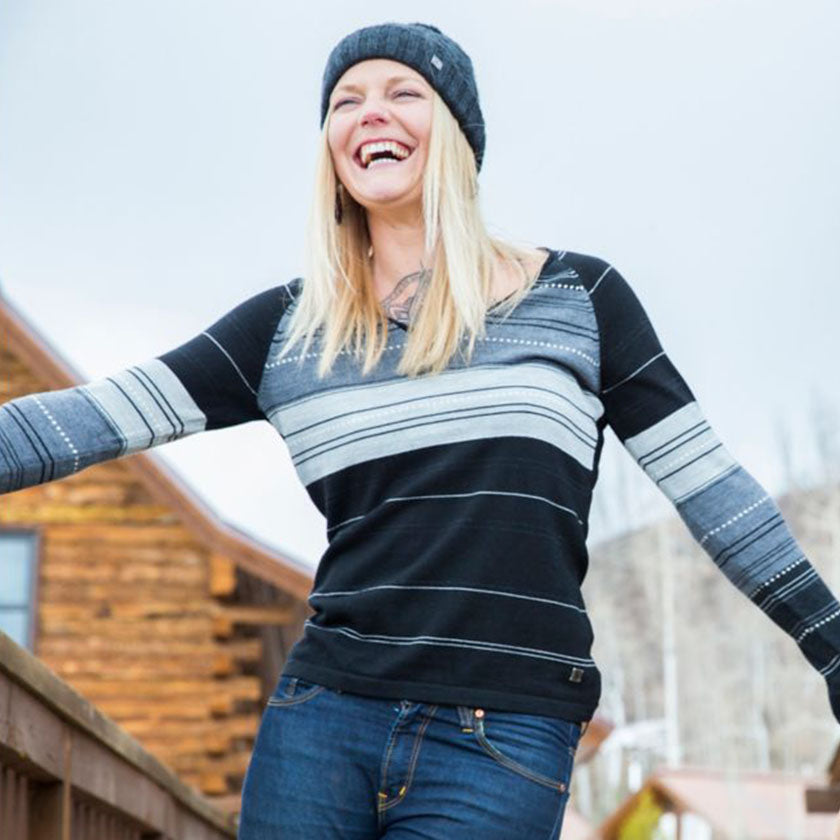 A blond woman in a Smartwool hat and sweater smiles in front of a wooded cabin. 