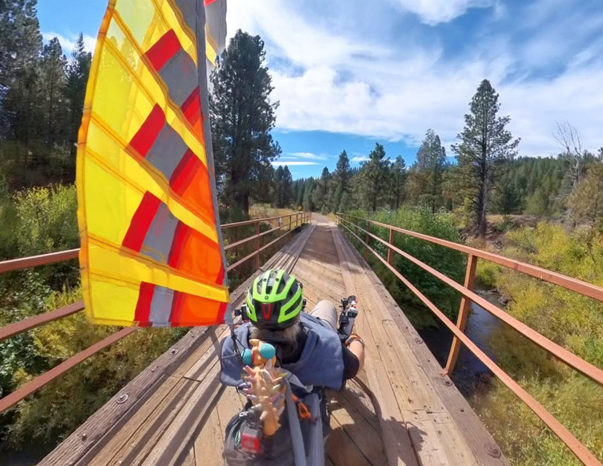 A person wearing a green helmet rides a recumbent trike with a large yellow, orange, and red reflective flag over a bridge, surrounded by trees and blue sky.