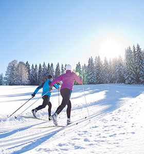 Two cross country skiers glide across the snow towards a tree line of pine trees covered in snow on a sunny day.