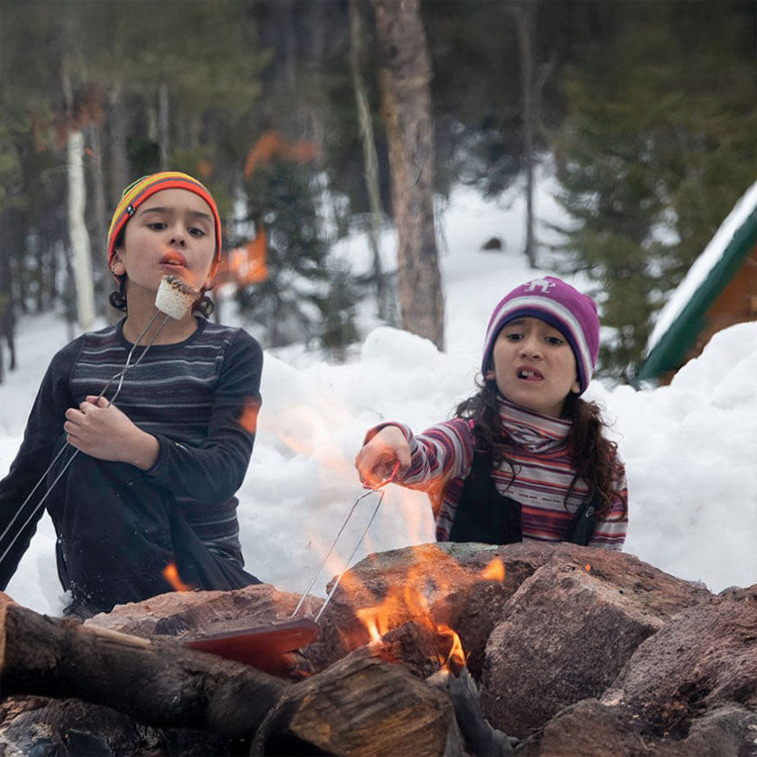 Two children in Smartwool clothing roast marshmallows over an outdoor fire surrounded by pine trees and snow.