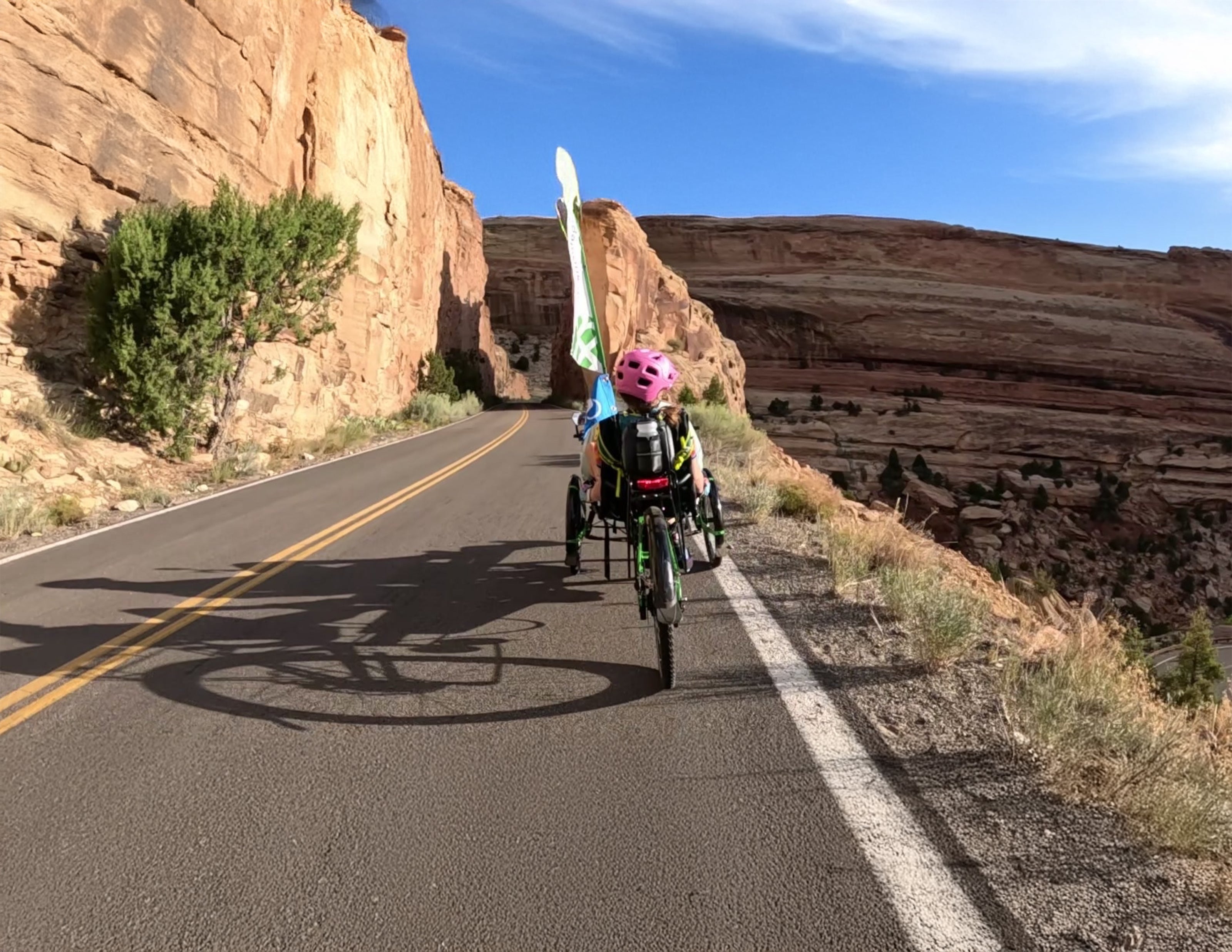 Person riding a recumbent trike on a desert road with rocky cliffs and clear sky.