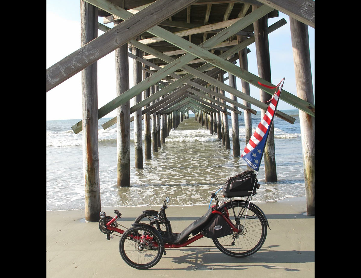 A red and black recumbent tricycle with a black saddlebag and a red, white, and blue flag, parked on a sandy beach under a wooden pier with the ocean in the background.