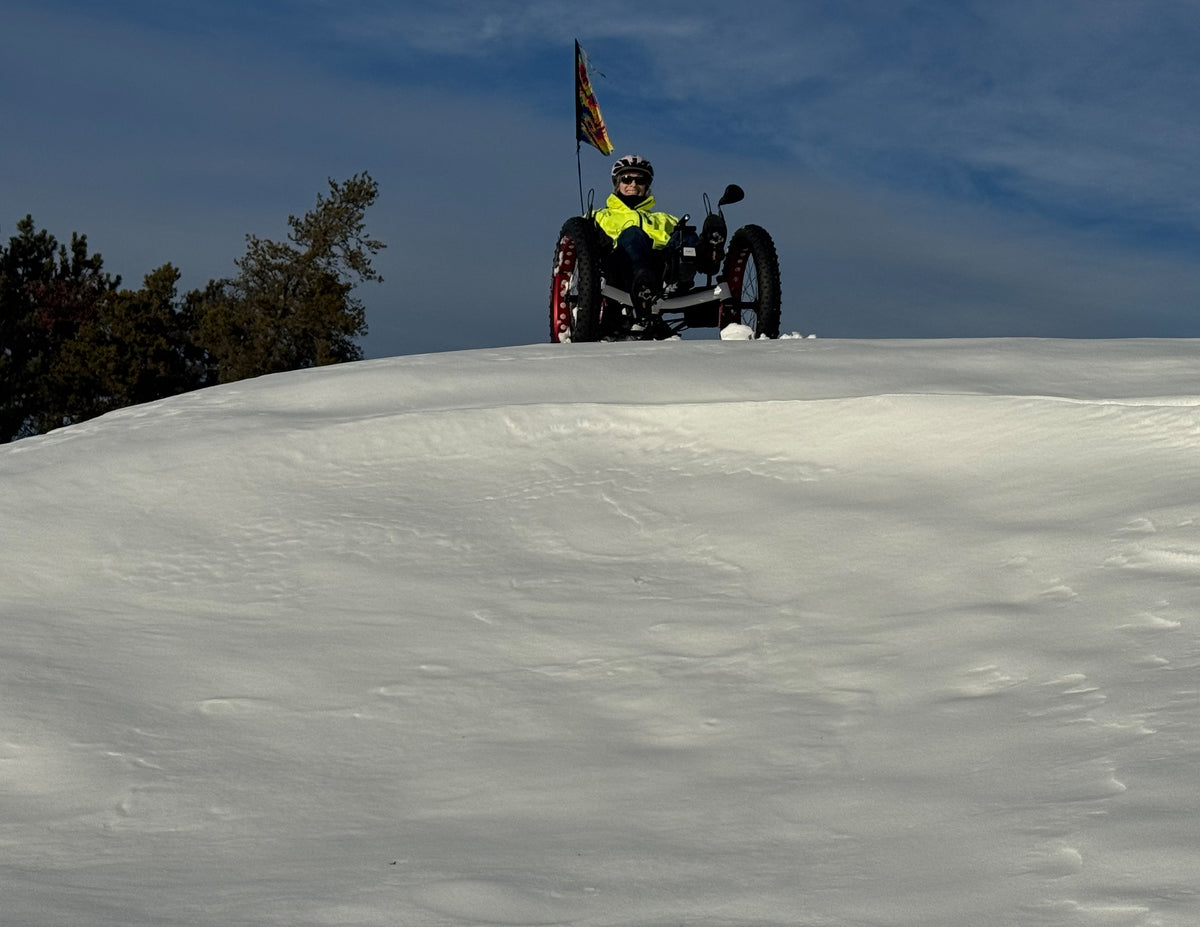 A person in a bright yellow jacket sits on a recumbent trike on a snowy hill under a blue sky, with trees visible on the left.