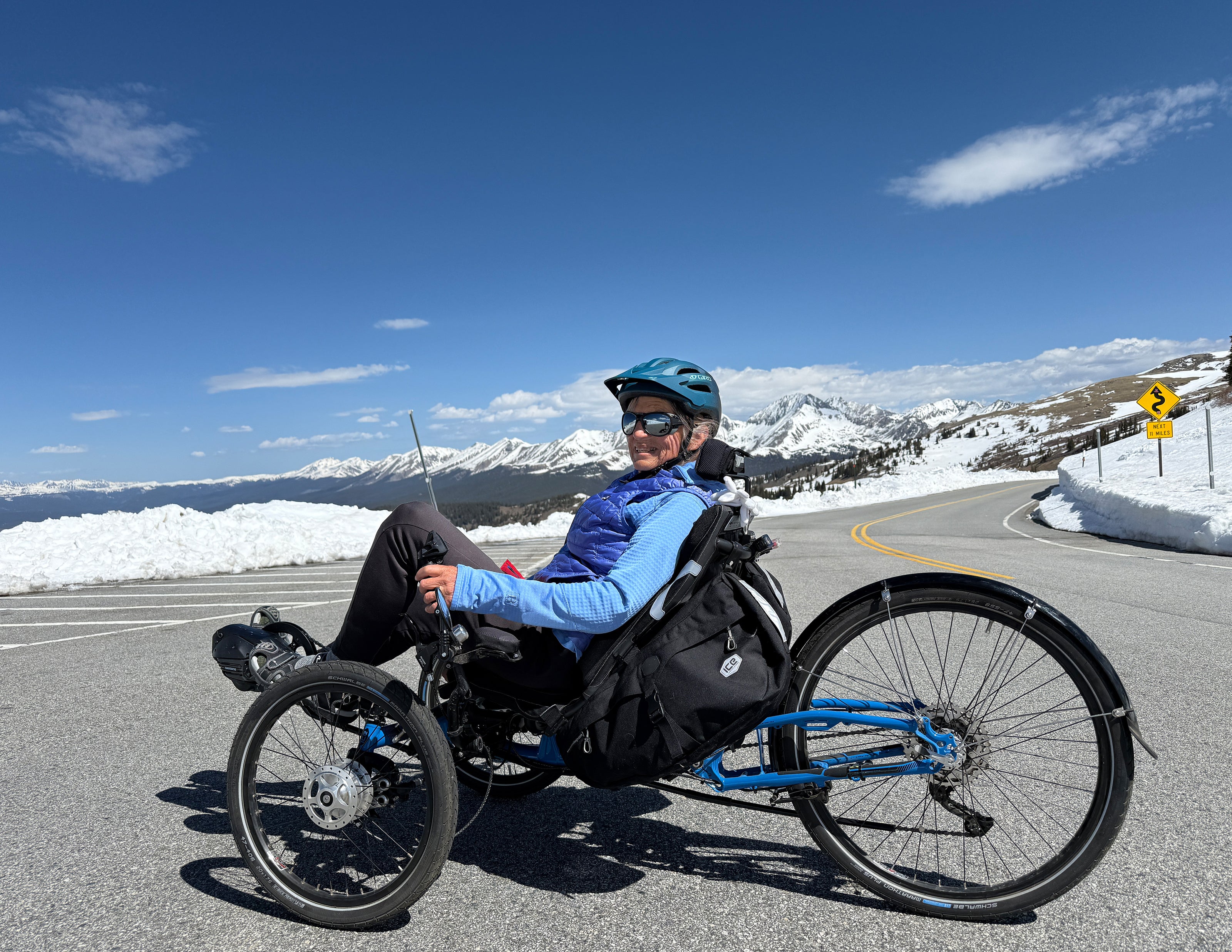 Person using a recumbent trike on a snowy mountain road with a clear blue sky.
