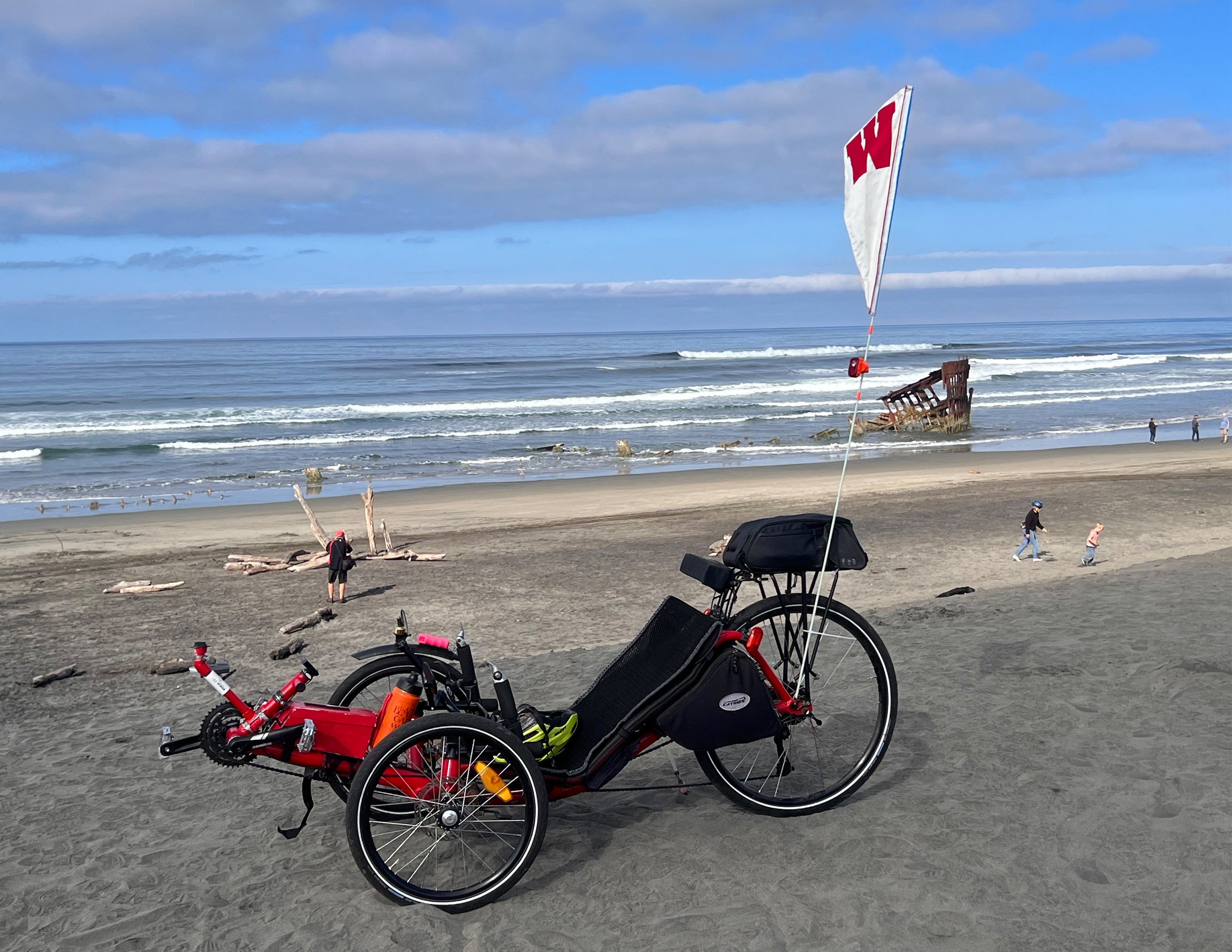 A recumbent trike, with a UW Wisconsin flag on it, on a sandy beach with an ocean view.