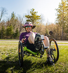 A woman in a purple top rides a green Catrike Pocket Recumbent Trike on a sunny day.