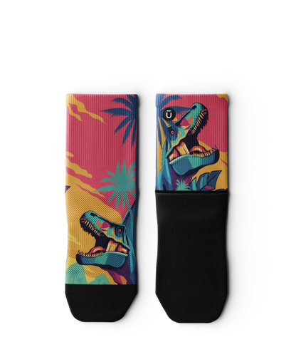 Multicolored  pair of socks laying flat with T-rex dinosaurs and palm trees on main part of sock, black soles. 