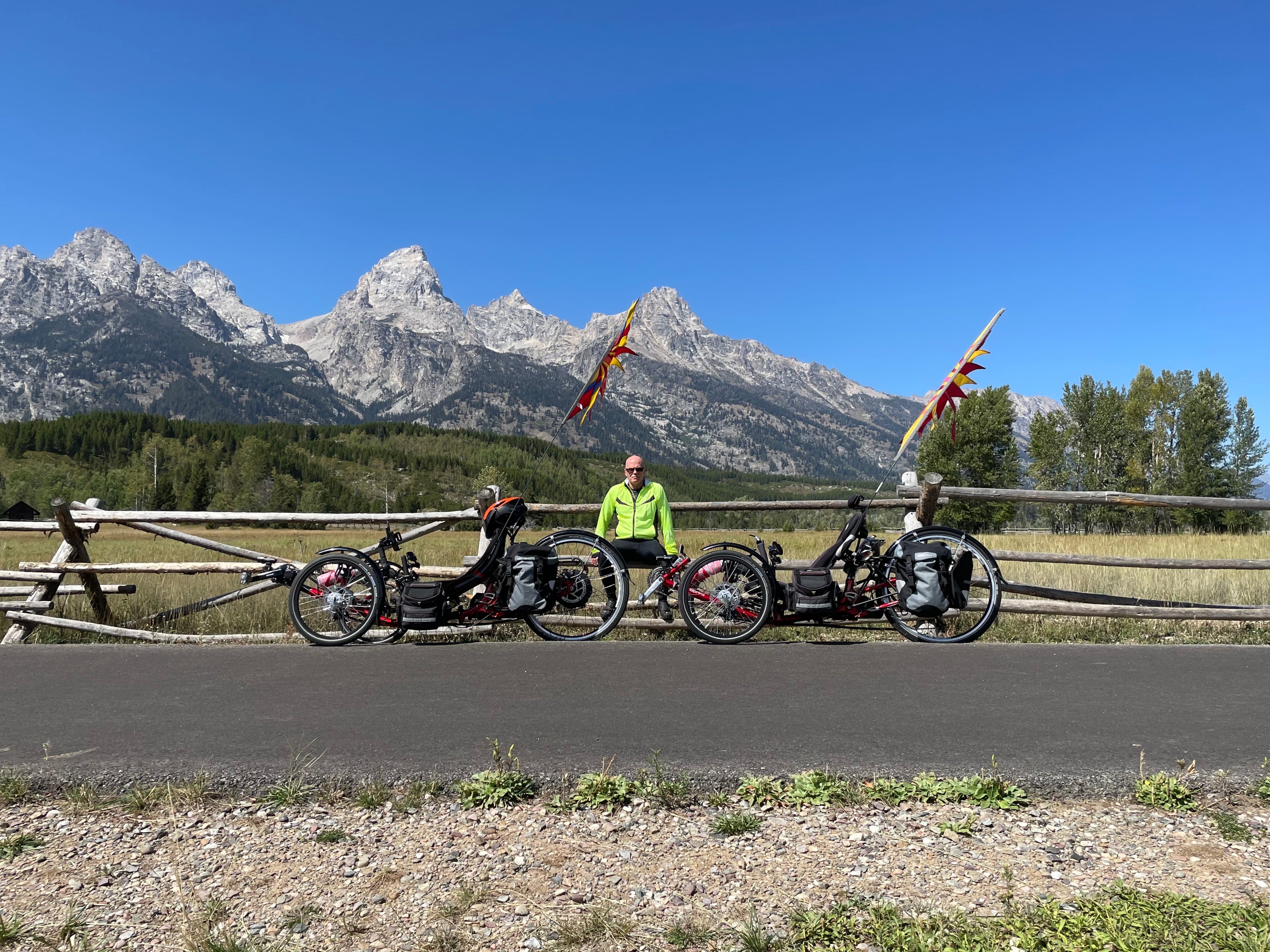 A rider poses with two recumbent tricycles in front of a scenic mountain backdrop.