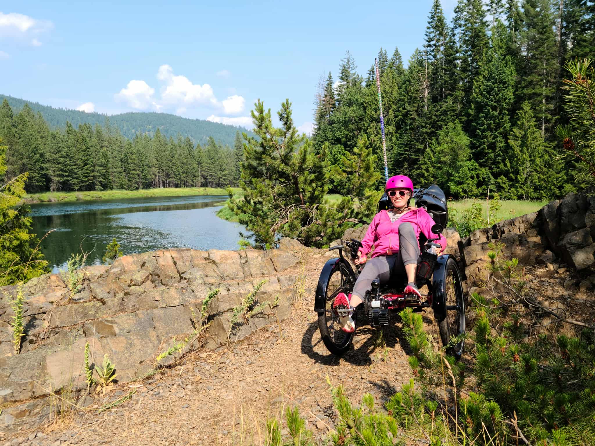 A person riding a recumbent trike on a trail near a lake with trees in the background.