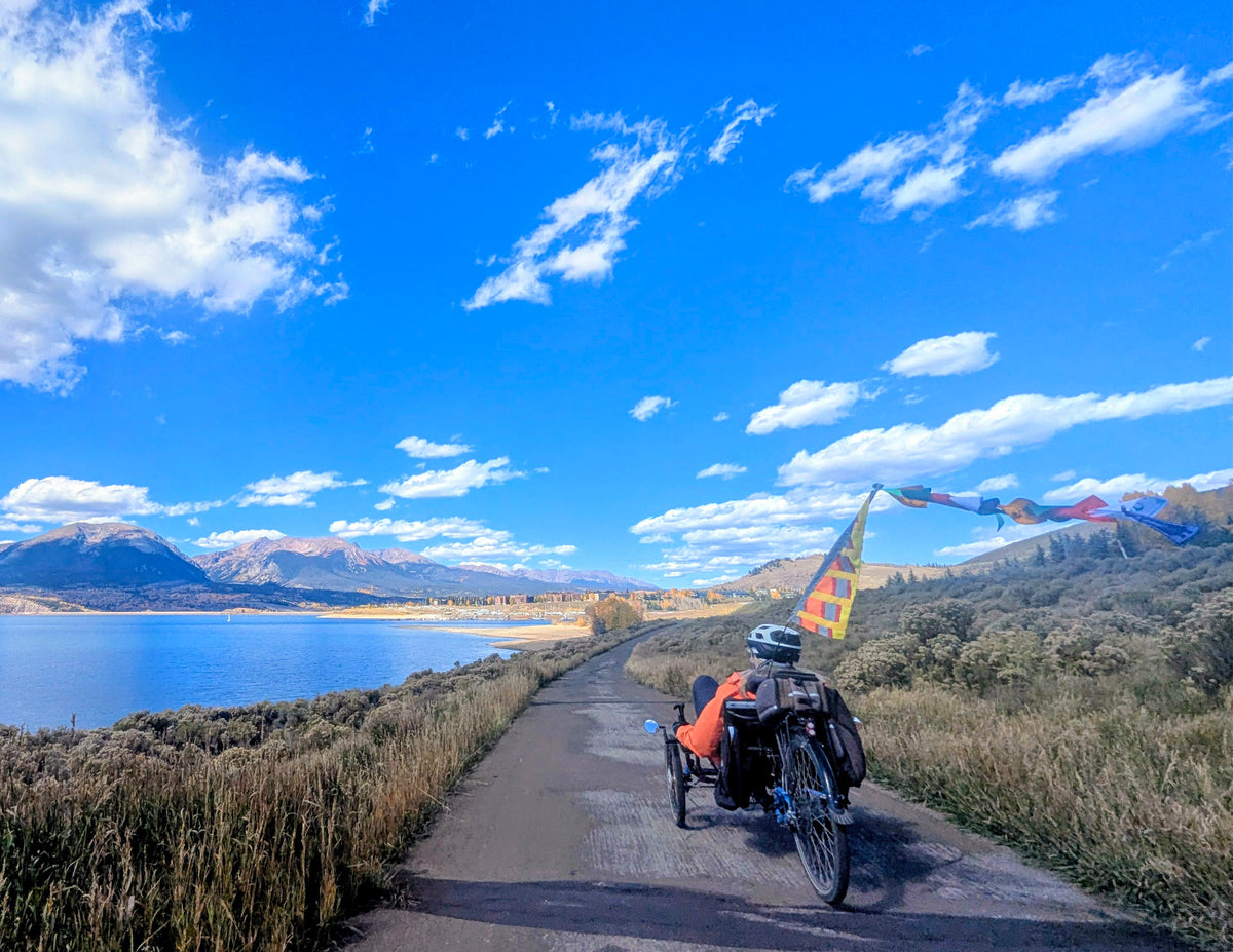 A person rides a recumbent tricycle along a paved path beside a lake, with mountains, blue sky, and clouds in the background. The tricycle is loaded with gear and has colorful flags attached.