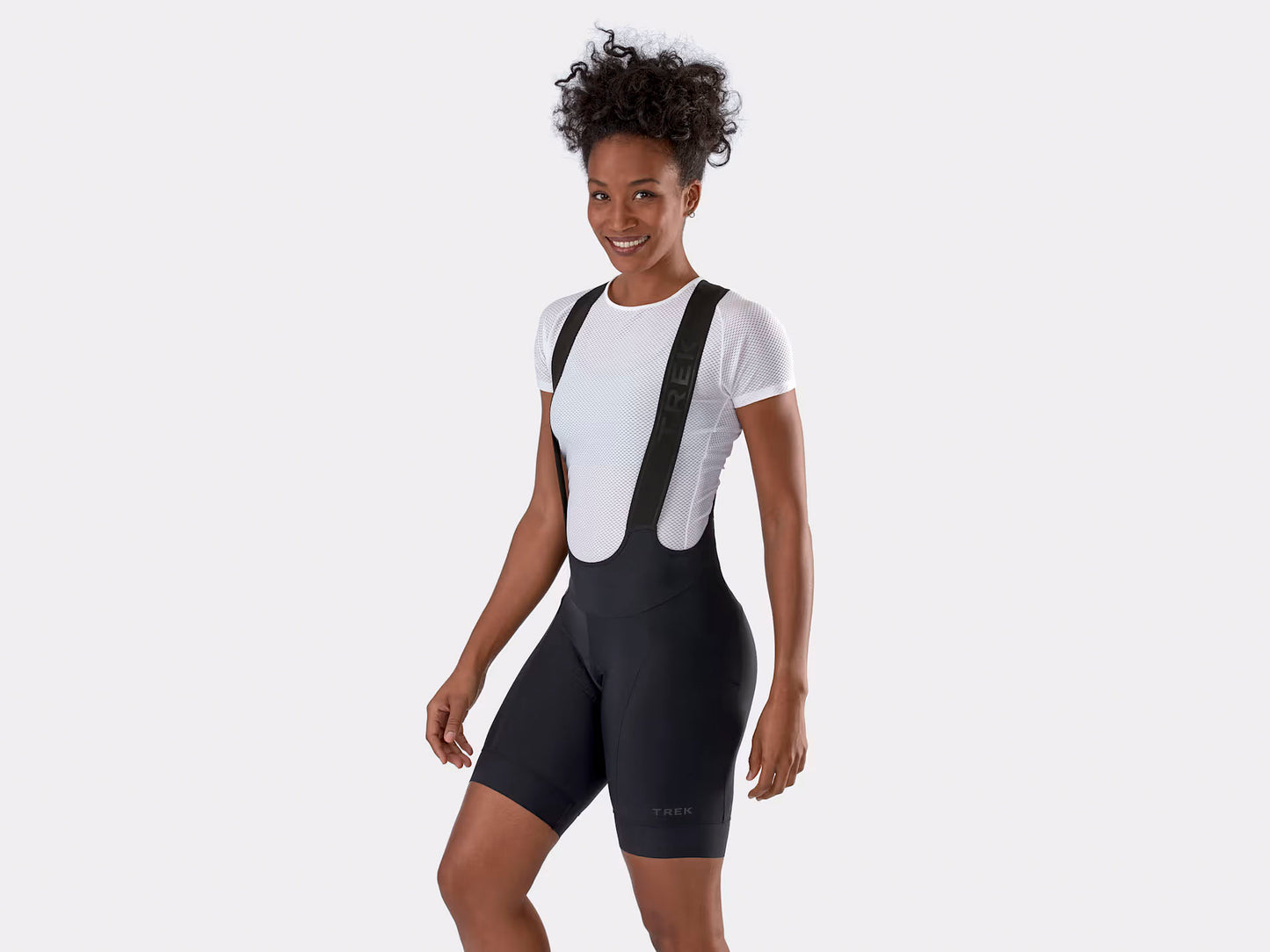 Studio front quarter view of a woman wearing black bib bicycle shorts over a white t-shirt.  Trek logo is embossed over cuff of left leg and along bib straps. 