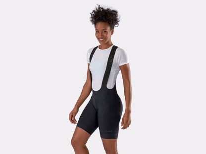 Studio front quarter view of a woman wearing black bib bicycle shorts over a white t-shirt.  Trek logo is embossed over cuff of left leg and along bib straps. 