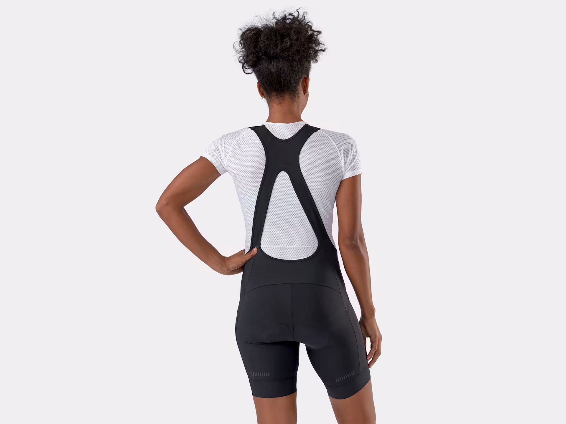 Studio back view of woman wearing black bib shorts over a white t-shirt.  Straps of bibs form an X across the back and there are reflective accents near the hem of the shorts.