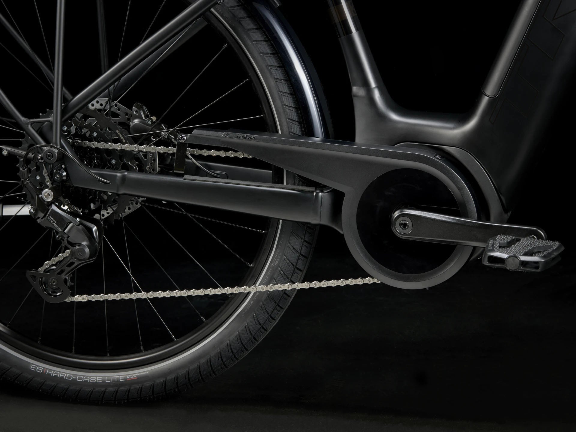 Side detail view of the drivetrain of the Trek Verve+ 3 Lowstep Gen 3 e-Bike in Black, shown on a black background. It includes fenders, lights, rear rack, suspension seatpost, and kickstand.