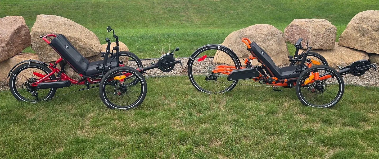 The Catrike Max in red and the Catrike Dumont in orange with e-assist kits installed against a grassy background with landscaping rocks.
