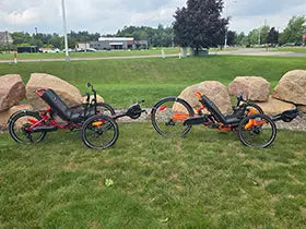 The Catrike Max in red and the Catrike Dumont in orange with e-assist kits installed against a grassy background with landscaping rocks.