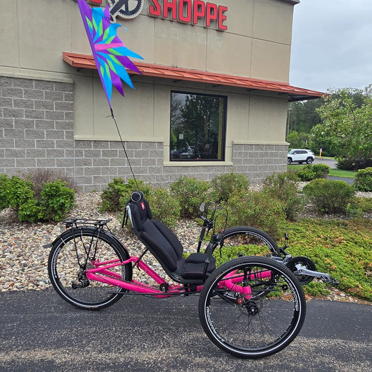 Side view of the Azub Used T-Tris AR Neon Pink Recumbent Trike with a colorful flag in an outdoor setting.