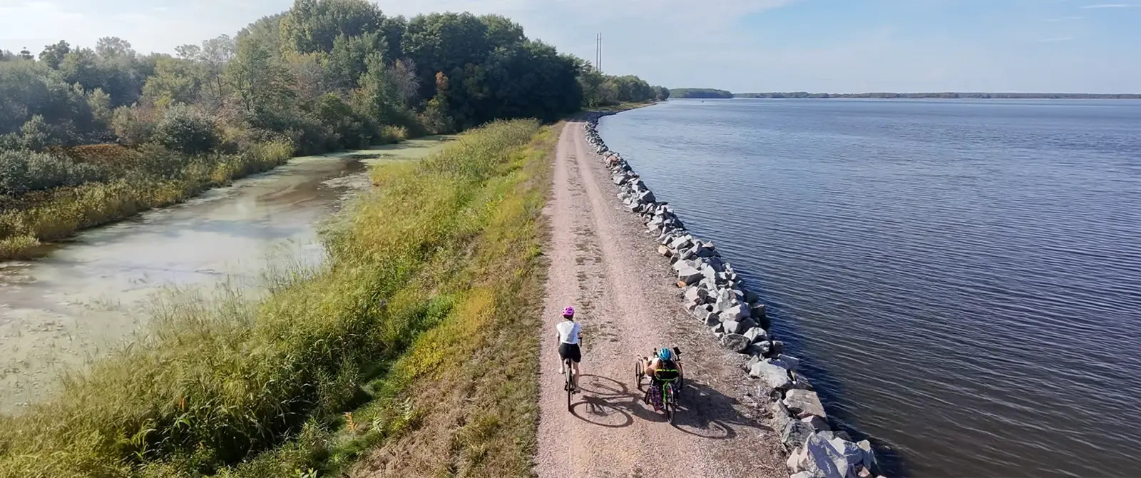 Two cyclists, a biker and a triker, on a path by the Wisconsin River in Stevens Point with trees in the background.