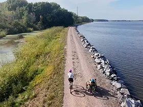 Two cyclists, a biker and a triker, on a path by the Wisconsin River in Stevens Point with trees in the background.