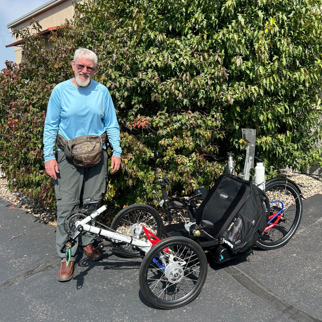 Man standing next to a recumbent trike with cargo bags in front of a bush outside the Hostel Shoppe.