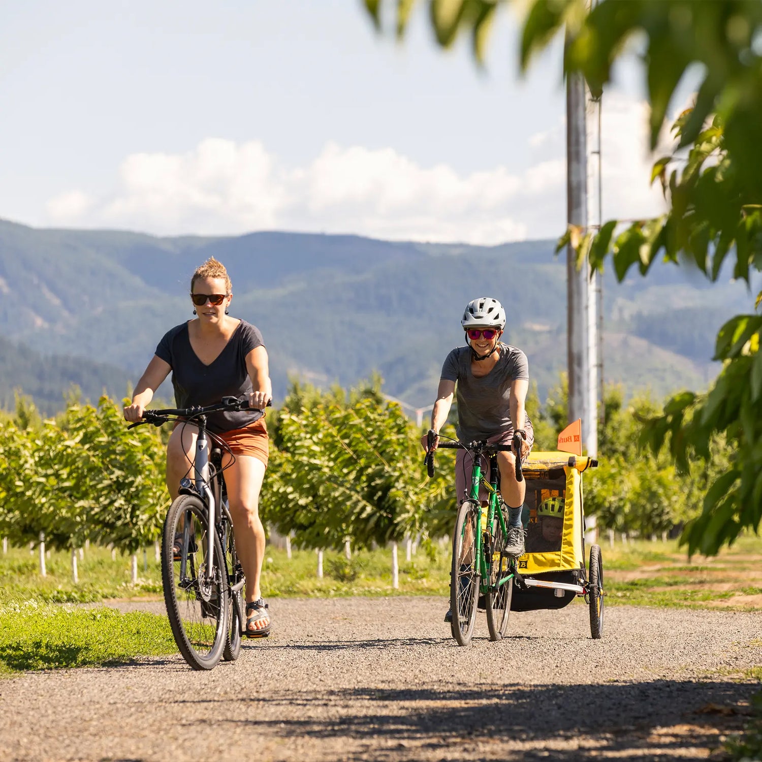 Two women cycle down a country road while one pulls a Burley child trailer on a sunny summer day.