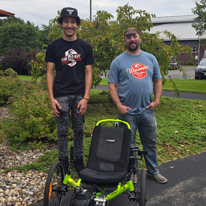 Two men in Pedal Point Rally t-shirts standing next to a green and black recumbent trike on a paved path with trees and a building in the background.