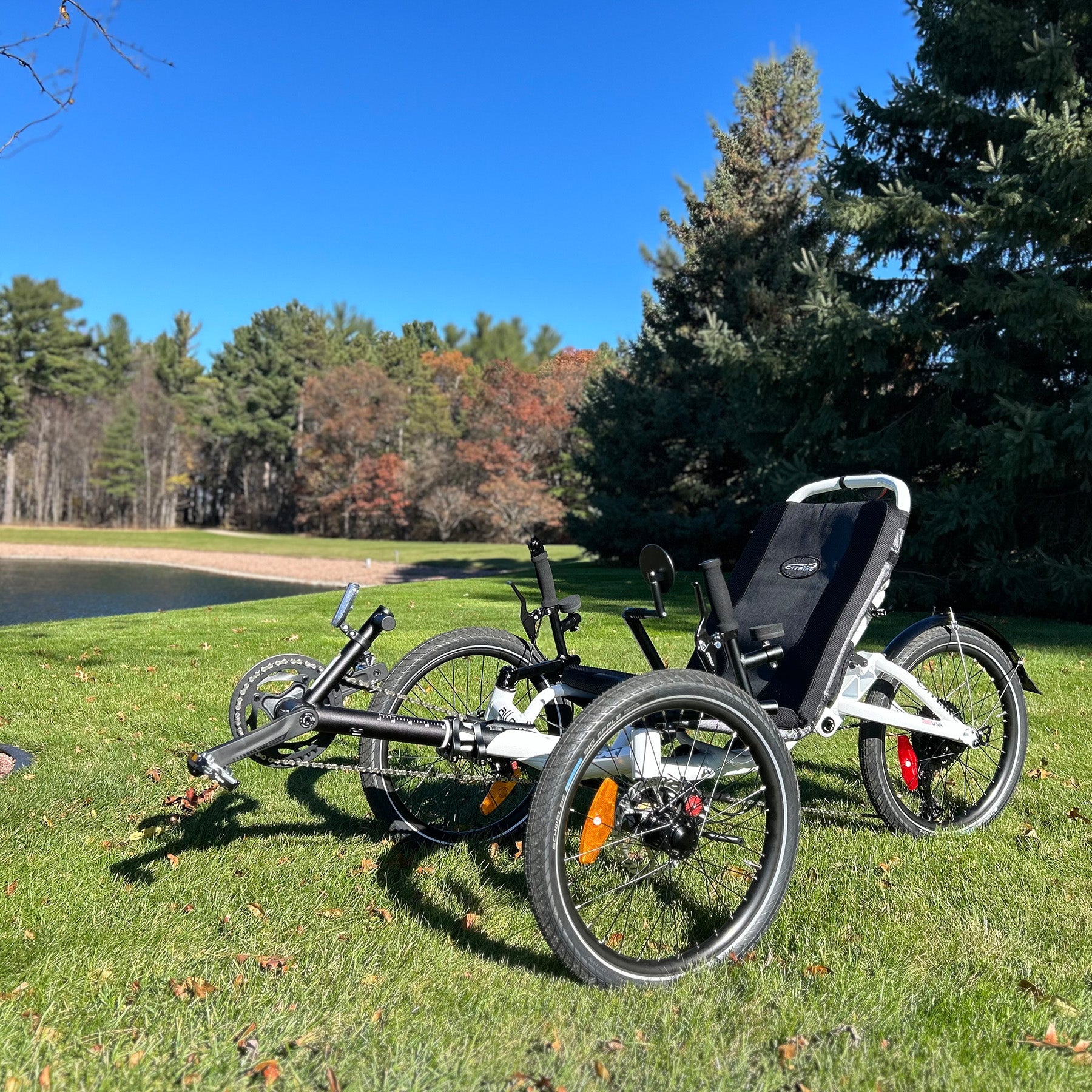 Three-wheeled recumbent trike on grass with trees and a lake in the background.