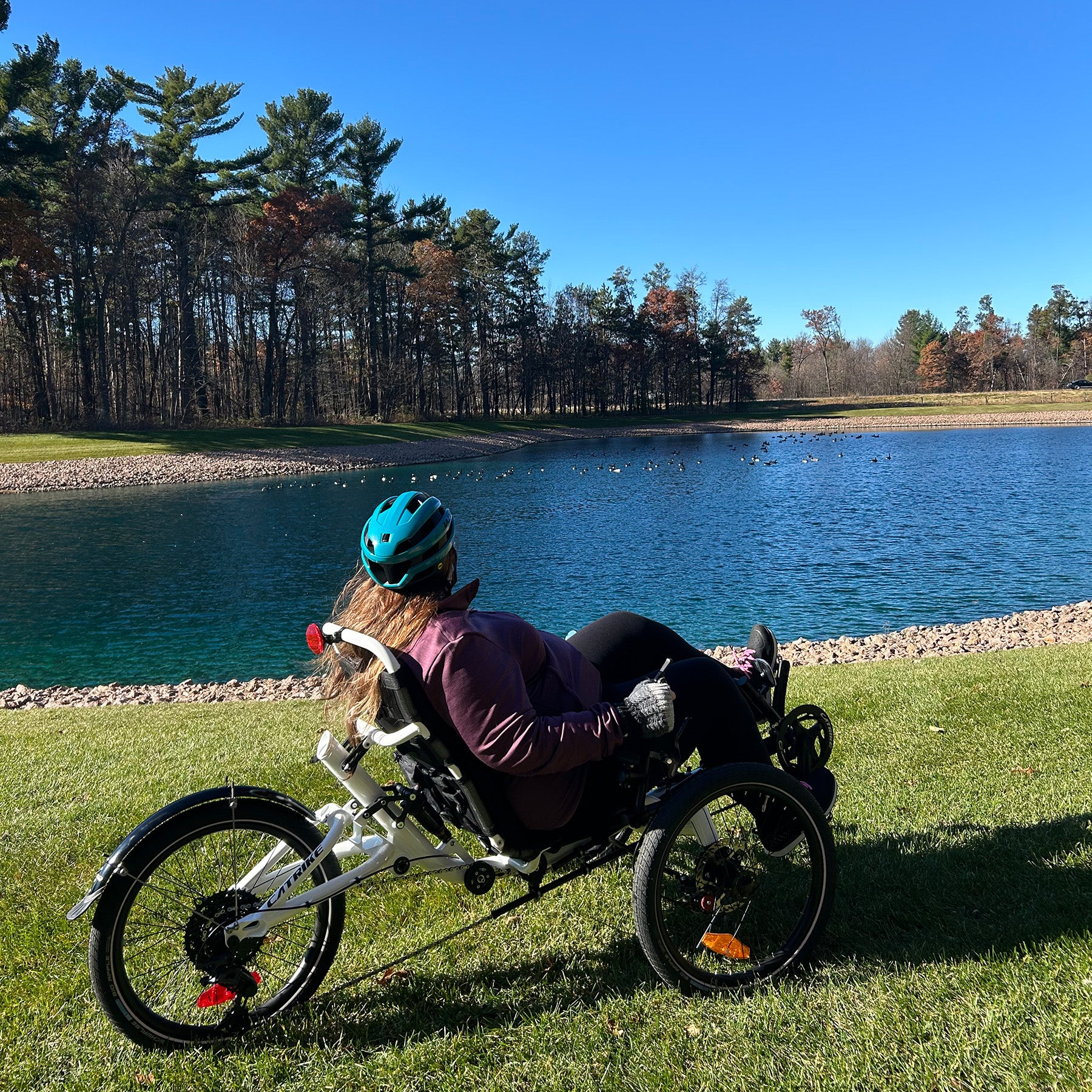 Person using a recumbent trike by a lake with trees in the background.