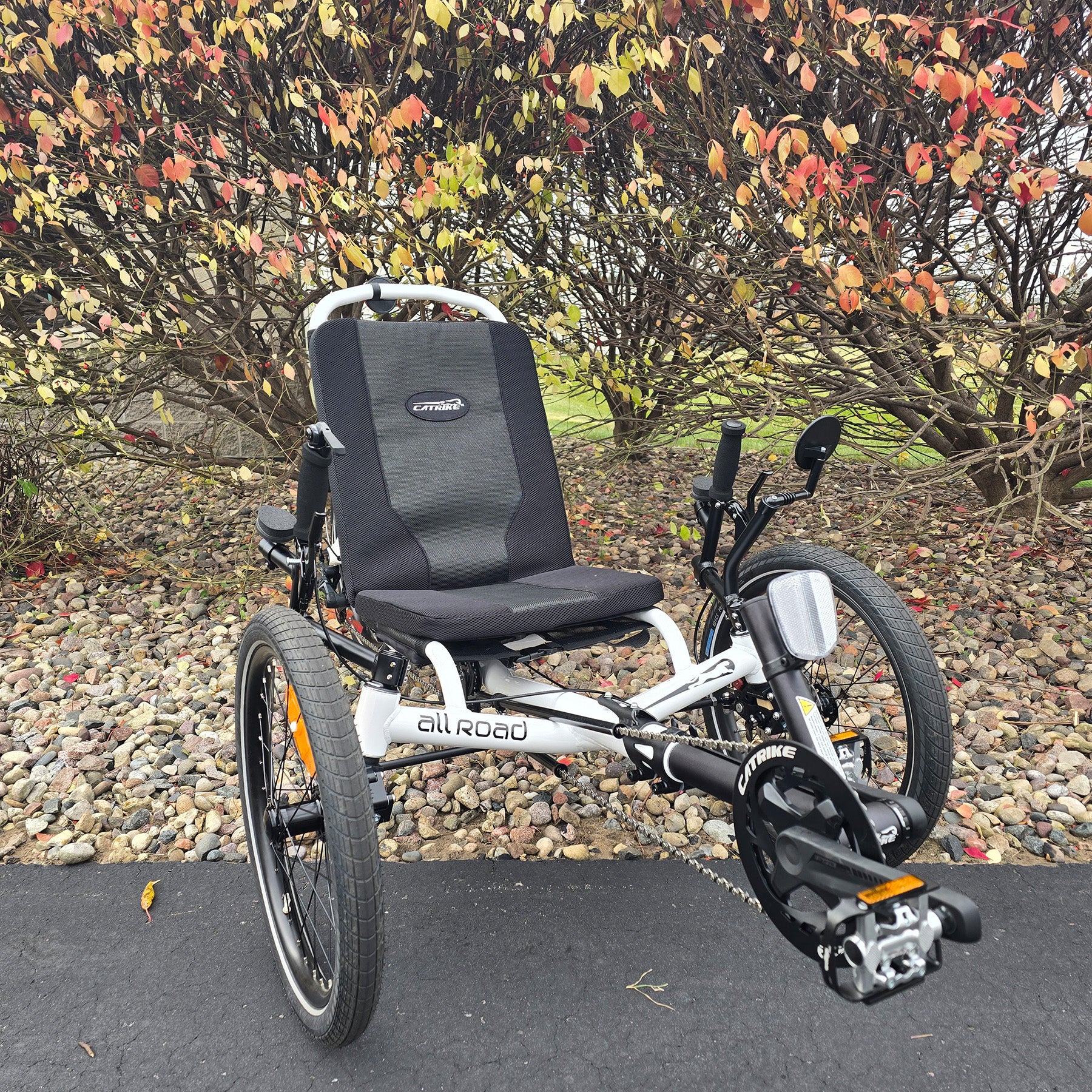 White recumbent trike with a black seat in an outdoor setting with trees and rocks.