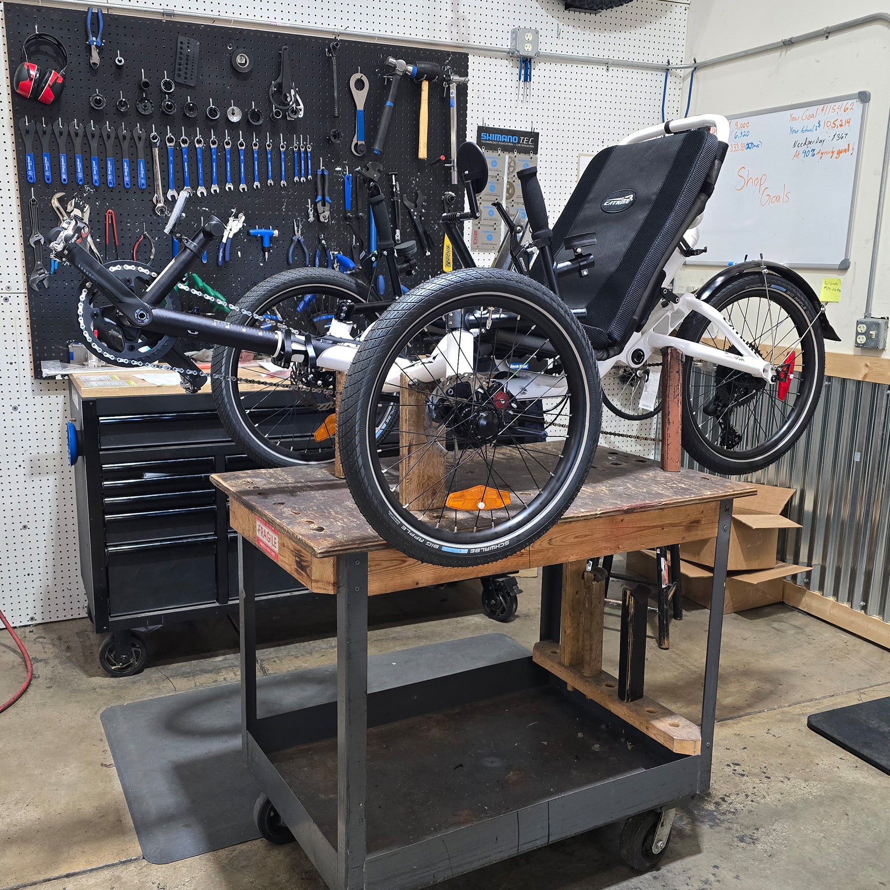 Recumbent trike on a workbench in a workshop with tools and equipment in the background.
