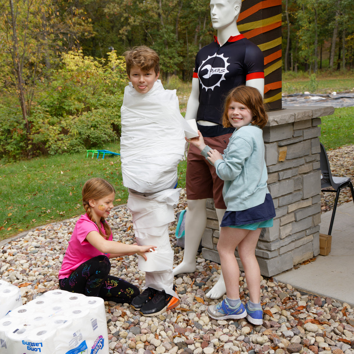 Three children are outdoors at the Green Goblin Get Together near a mannequin wearing a black and red jersey with a circular white logo. One child is wrapped in white toilet paper, one is kneeling and applying more paper, and one is standing beside the mannequin.