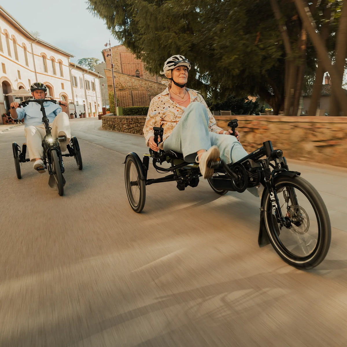 A man and a woman ride the Hase Kettwiesel One Plus Enviolo Bosch Performance CX Pale Green Recumbent Trike in a city setting.