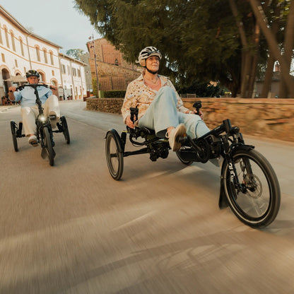 A man and a woman ride the Hase Kettwiesel One Plus Enviolo Bosch Performance CX Pale Green Recumbent Trike in a city setting.
