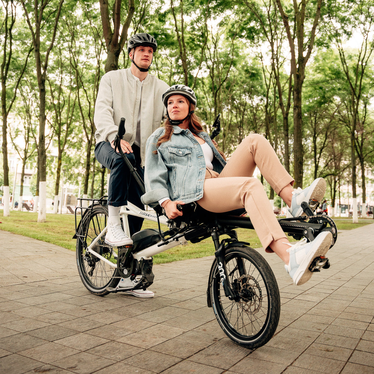 A man and woman ride a Hase Pino EP6 Cargo Grey Beige Tandem Recumbent Bike in a tree filled city setting.