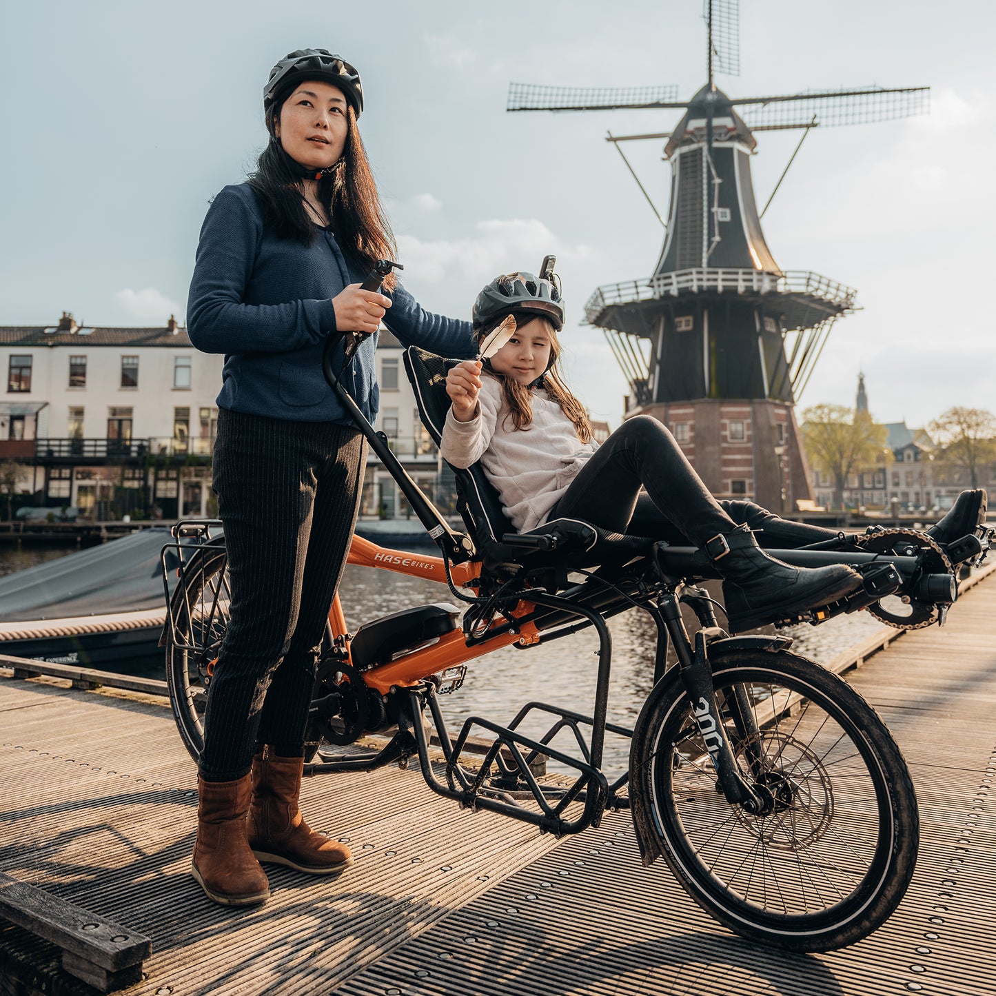 A woman and child ride a Hase Pino EP6 Cargo Grey Beige Tandem Recumbent Bike along a waterfront setting near a windmill.