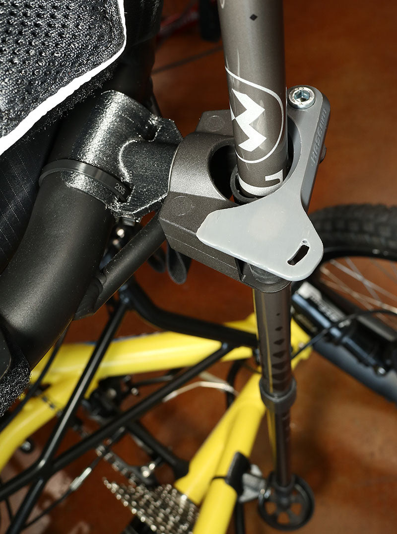Image of the top portion of a Hase Walking Stick Mount, with a walking stick attached, installed onto the seat frame of a yellow recumbent trike. 