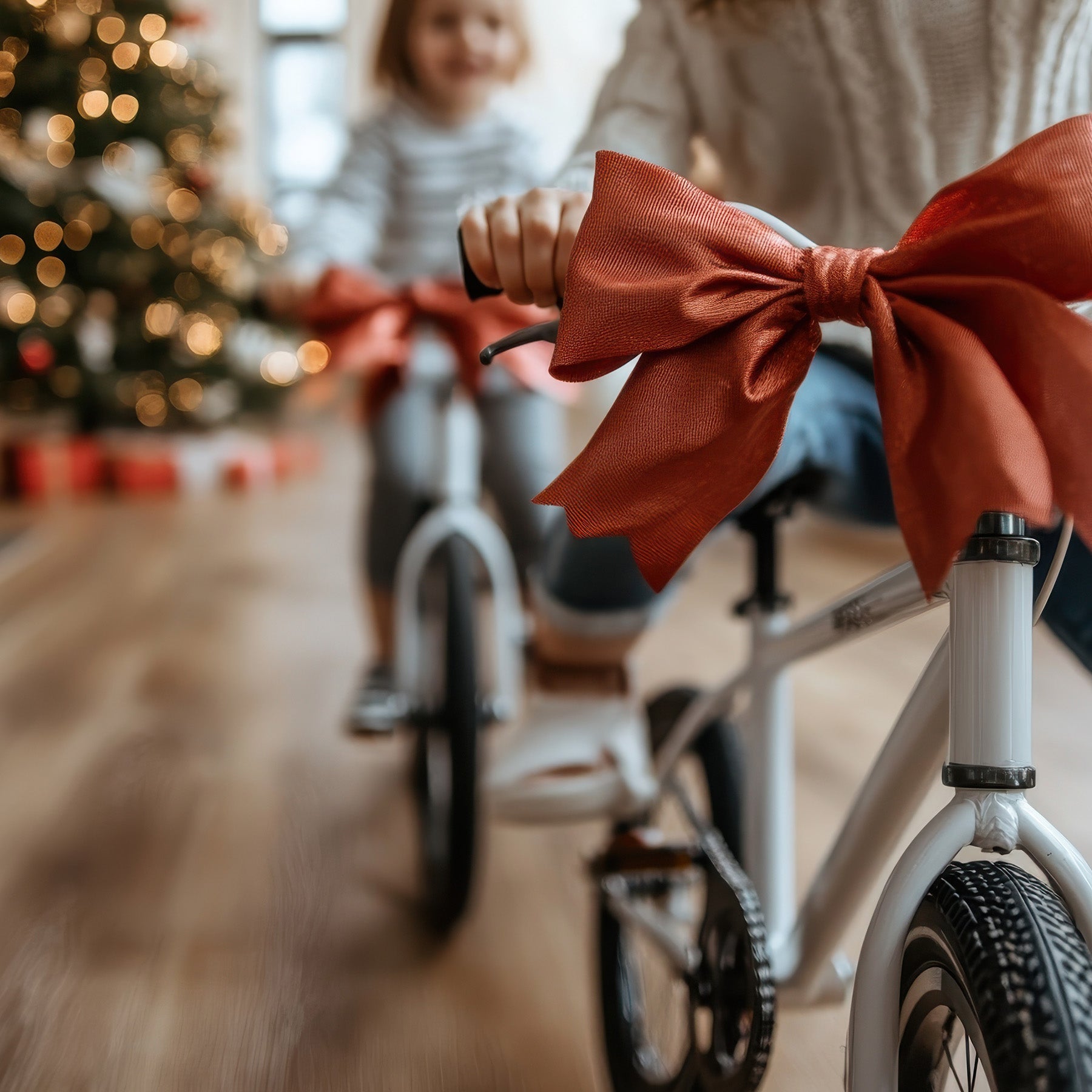 Two girls ride bikes with bows in a living room with a Christmas tree in the background.