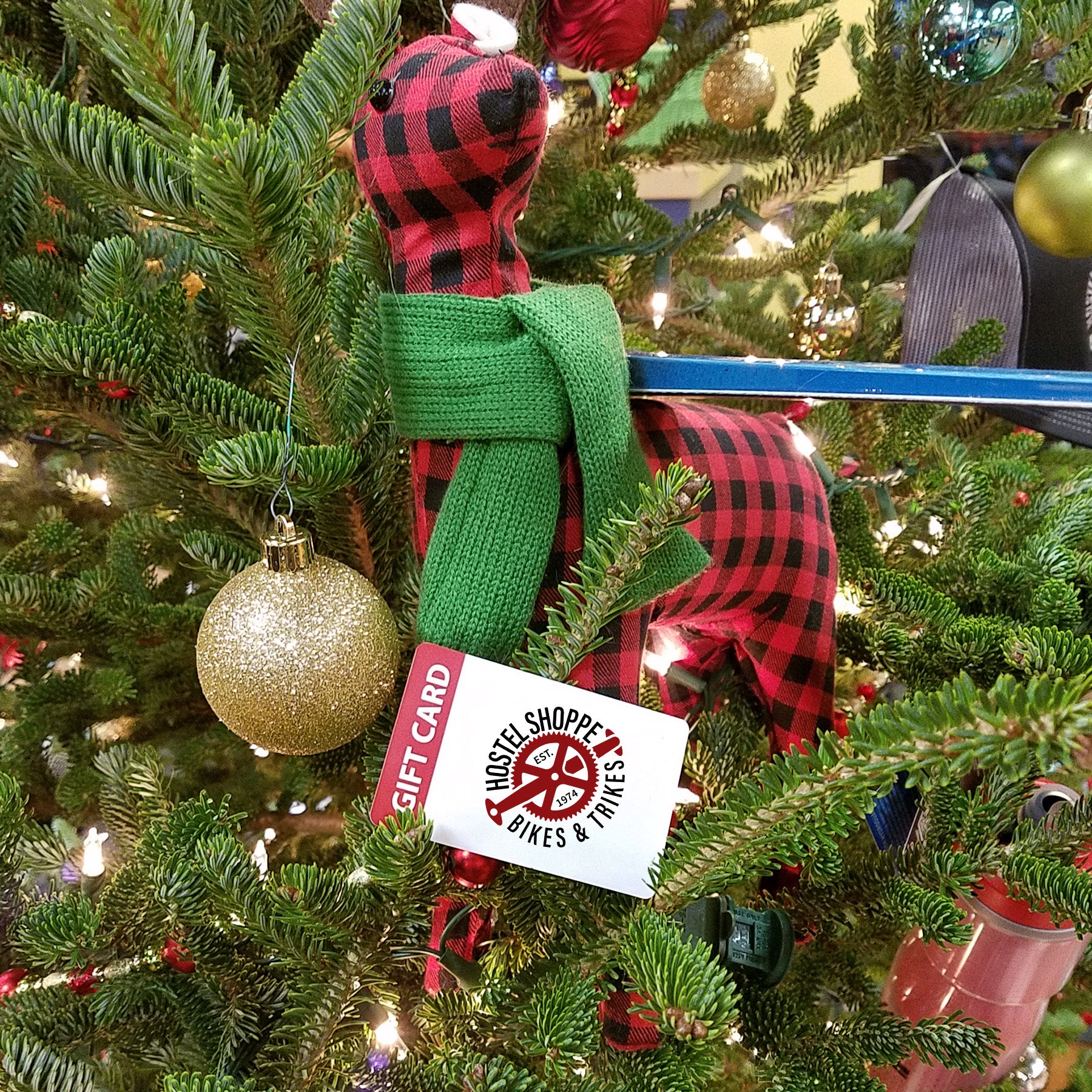 A Hostel Shoppe Gift Card is nestled in a Christmas tree under a plaid reindeer wearing a green scarf.