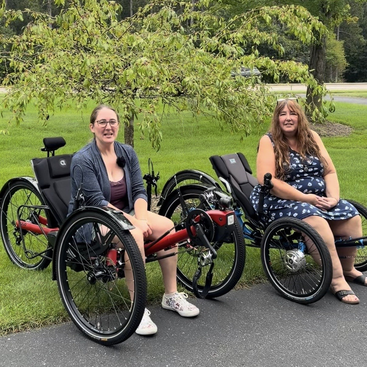 Two women sit on recumbent trikes outdoors on a paved path under a tree with green grass behind them.