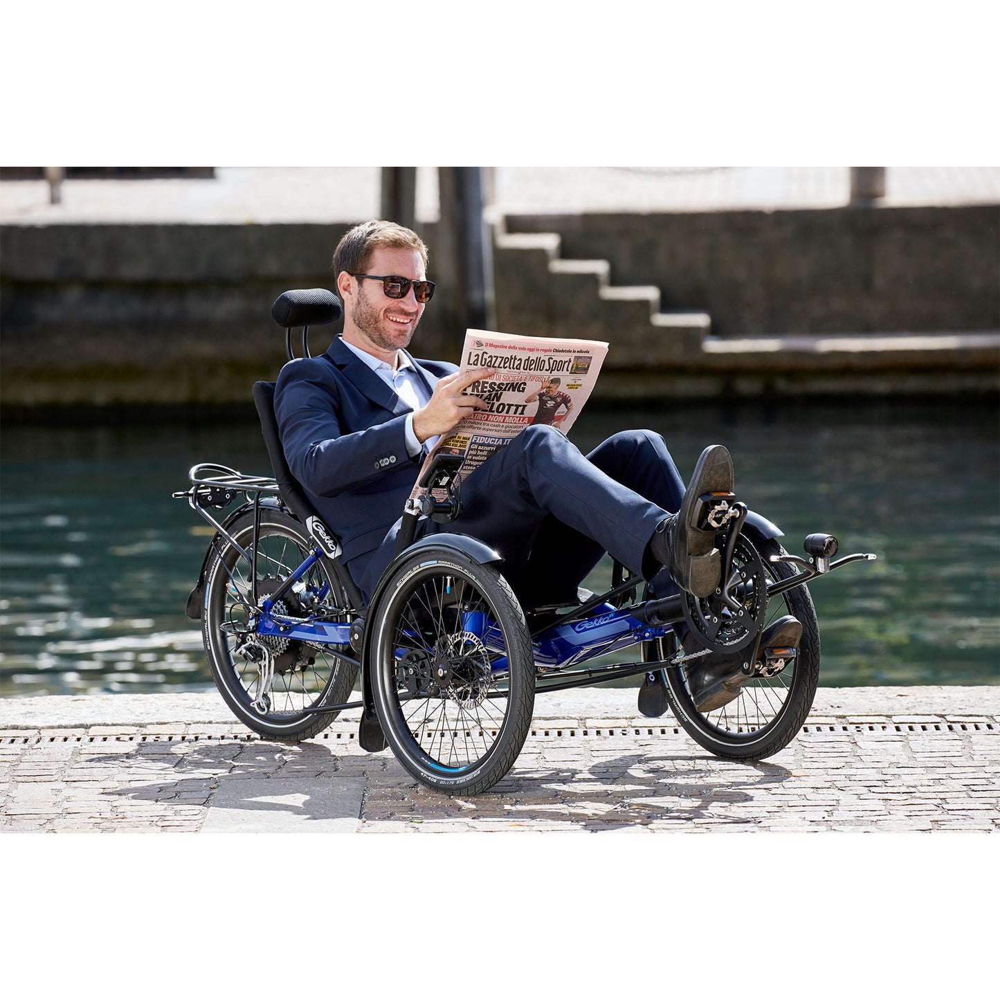 Man sitting on the HP Velotechnik Gekko fx 20 Sport Recumbent Trike in Deep Blue reading a newspaper alongside a river in the city.