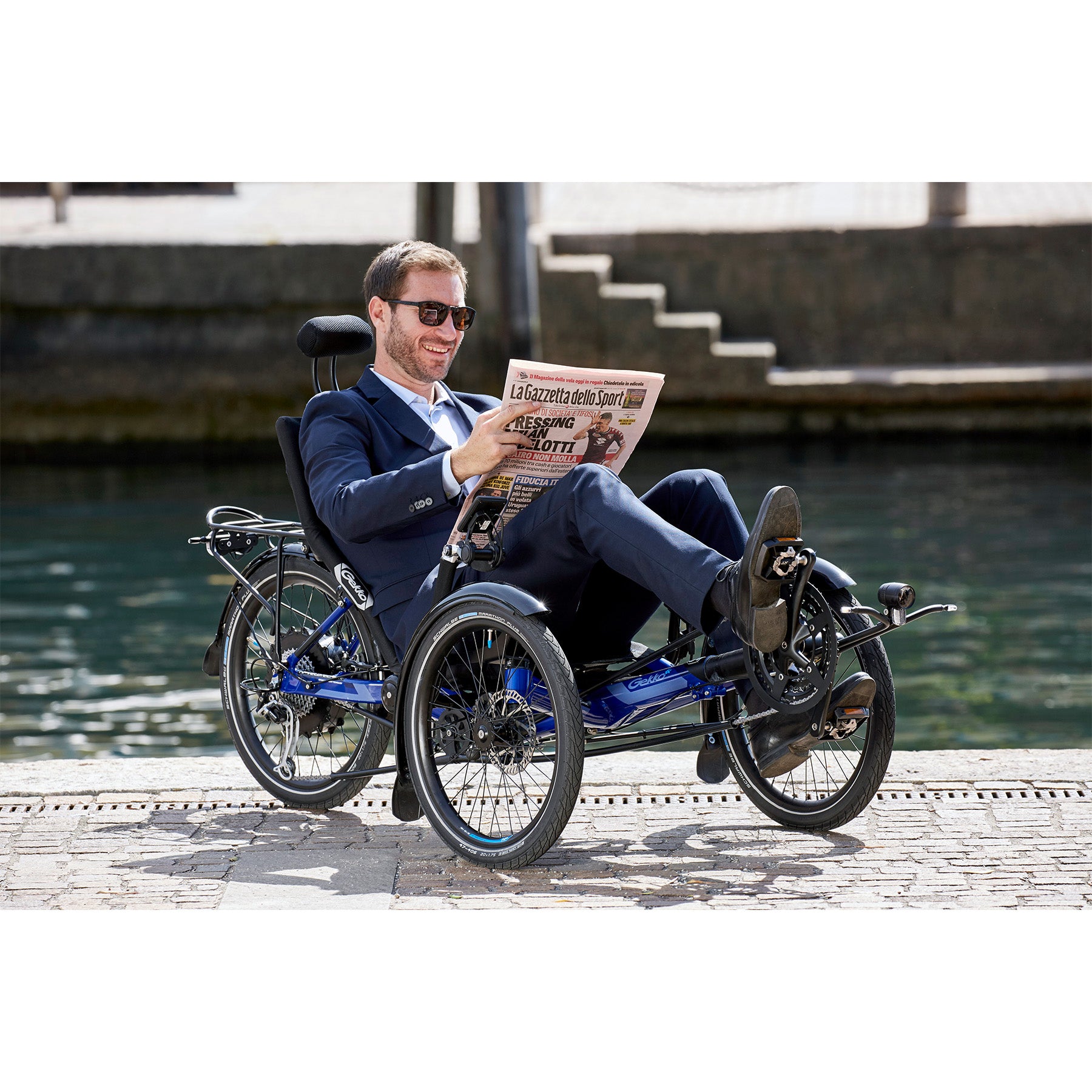 Man sitting on the HP Velotechnik Gekko fx 20 Sport Recumbent Trike in Deep Blue reading a newspaper alongside a river in the city.