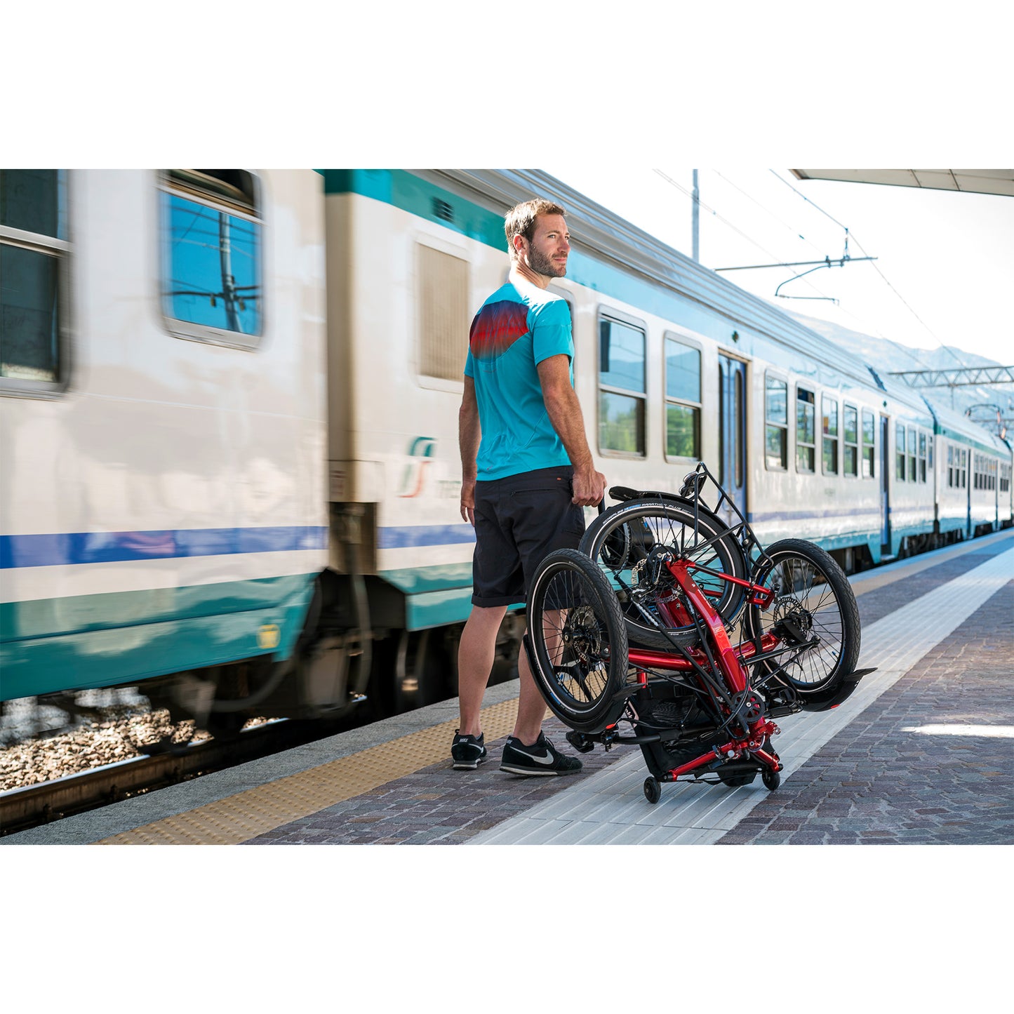 Man on a train platform with a folded HP Velotechnik Gekko fx 20 Sport Recumbent Trike in Magma Red.