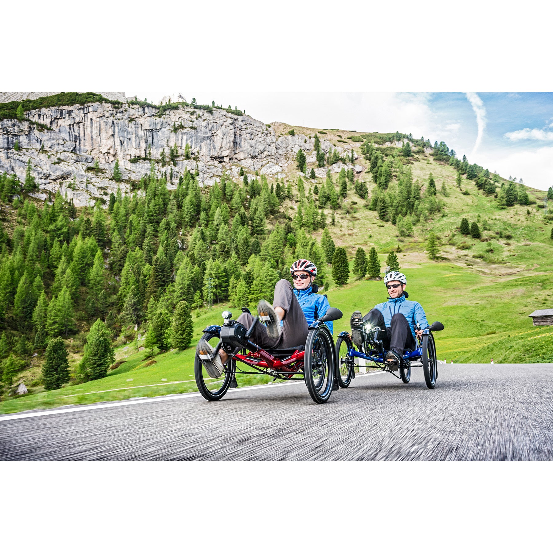 Two people riding recumbent trikes on a mountain road with greenery and cliffs in the background.