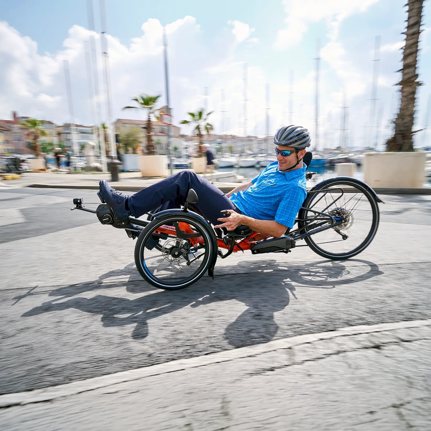 A man riding an HP Velotechnik Scorpion fs 26 recumbent trike with electric assist on a sunny day with palm trees in the background of a cityscape by the shore.