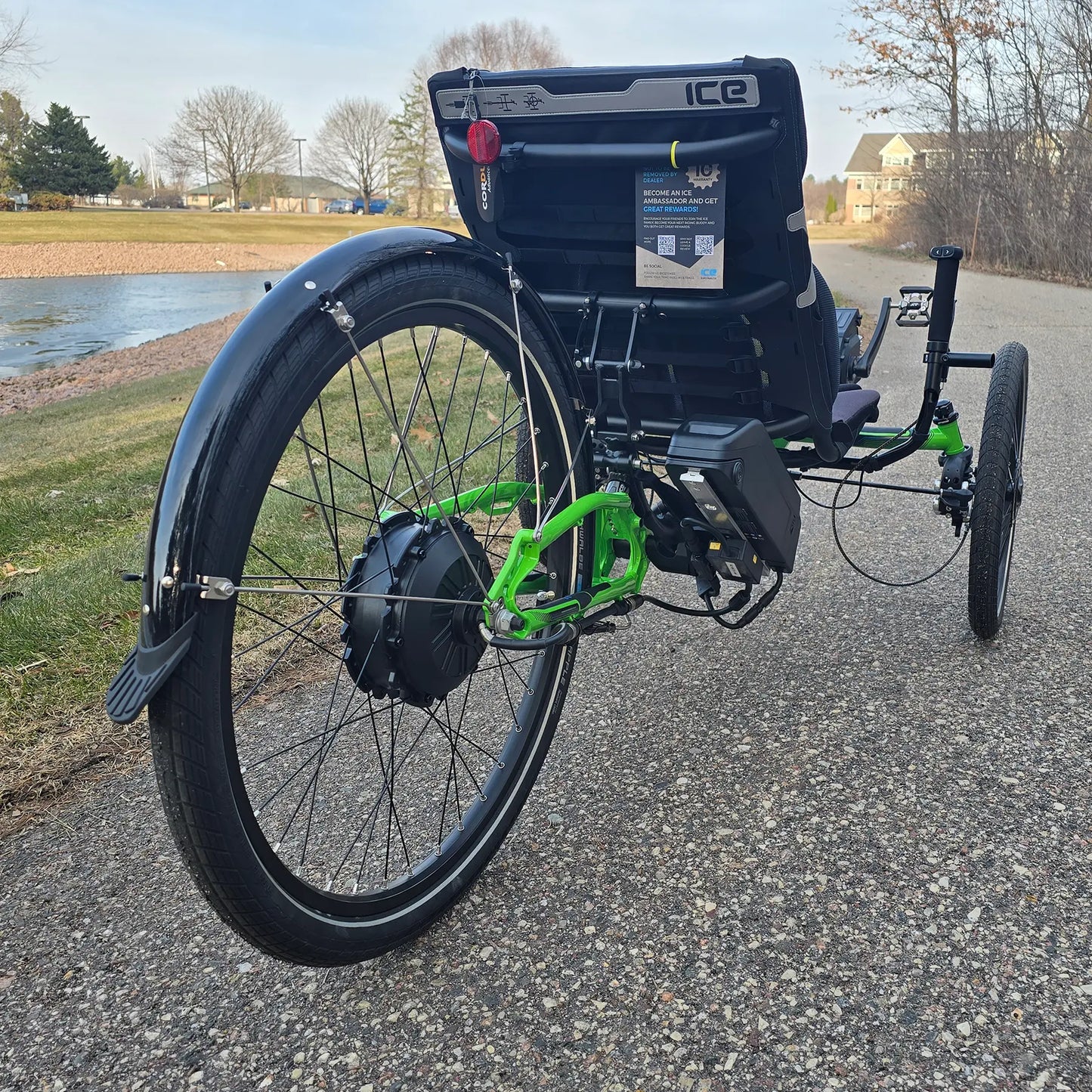 Rear view of the ICE Adventure HD 26 FS PERS Chainless Kiwi Green Recumbent Trike on a bike path near a melting pond with trees in the background.