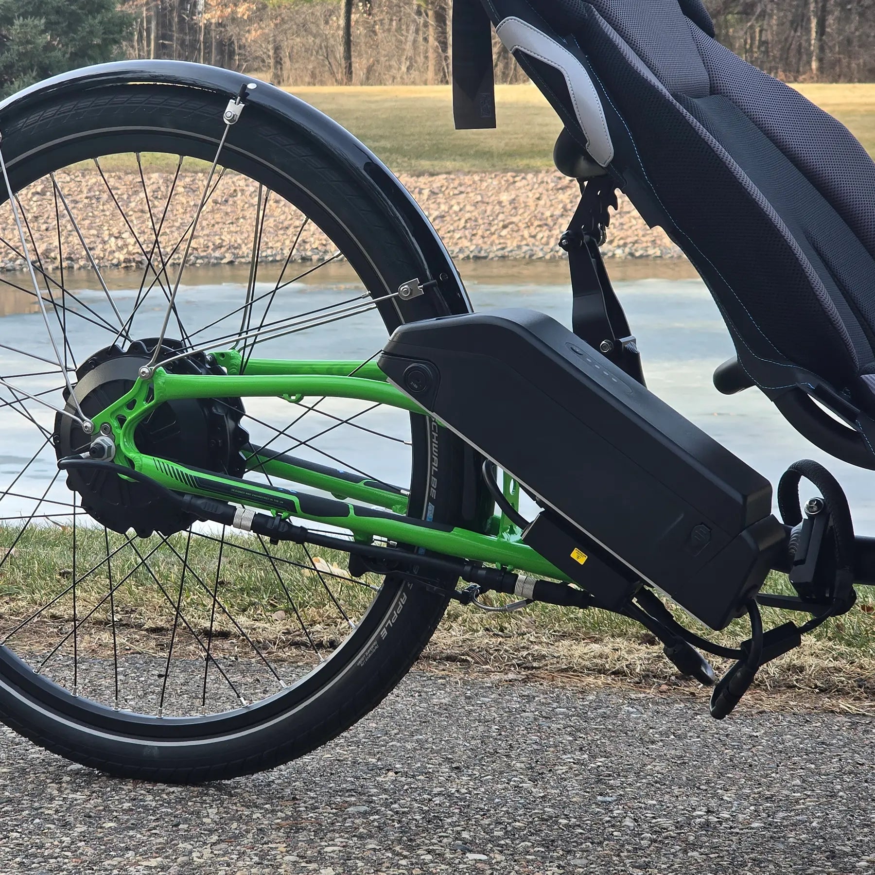 Closeup view of the rear wheel and b ttery on the ICE Adventure HD 26 FS PERS Chainless Kiwi Green Recumbent Trike on a bike path near a melting pond.