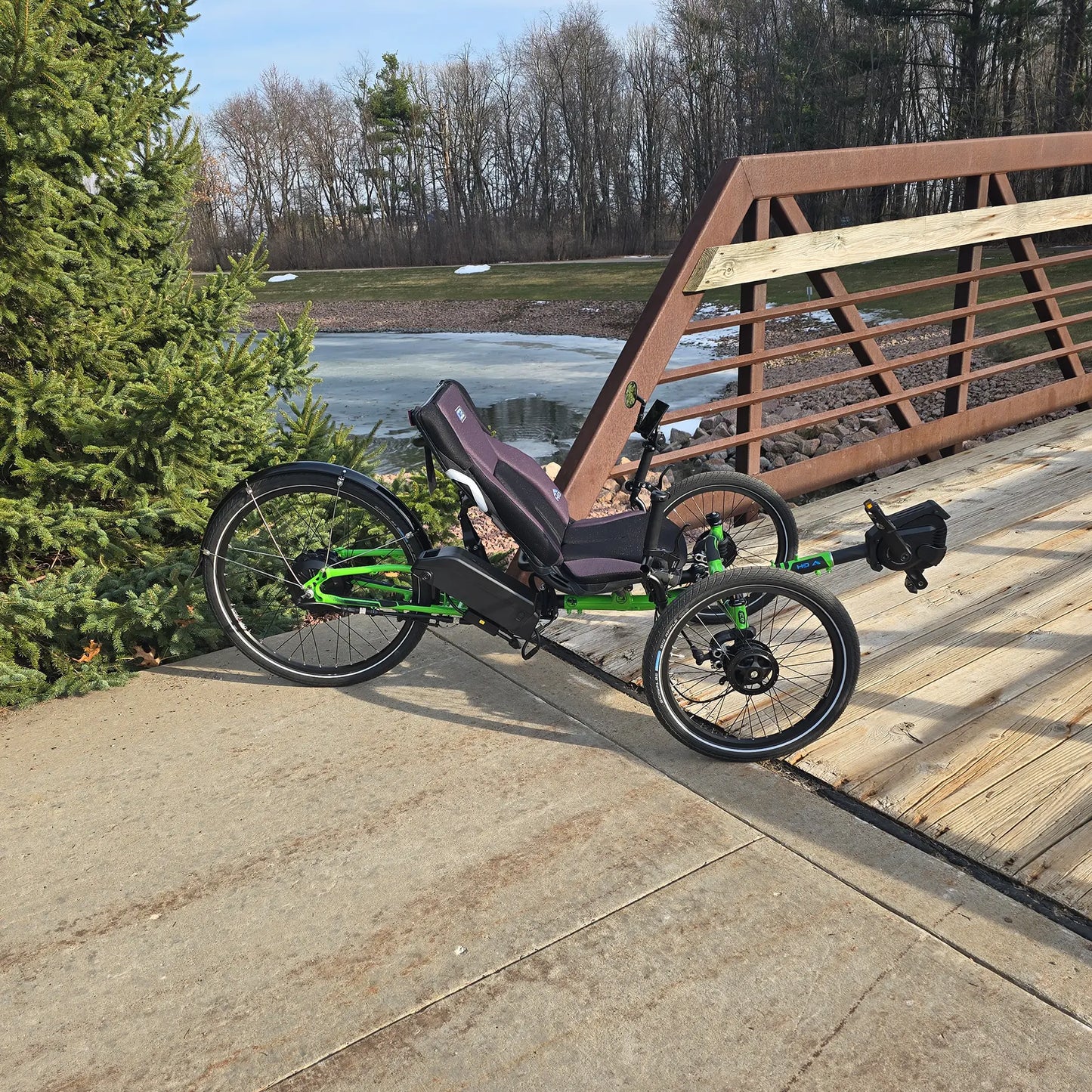 Side view of the ICE Adventure HD 26 FS PERS Chainless Kiwi Green Recumbent Trike on a bike path by a bridge near a melting pond with trees in the background.