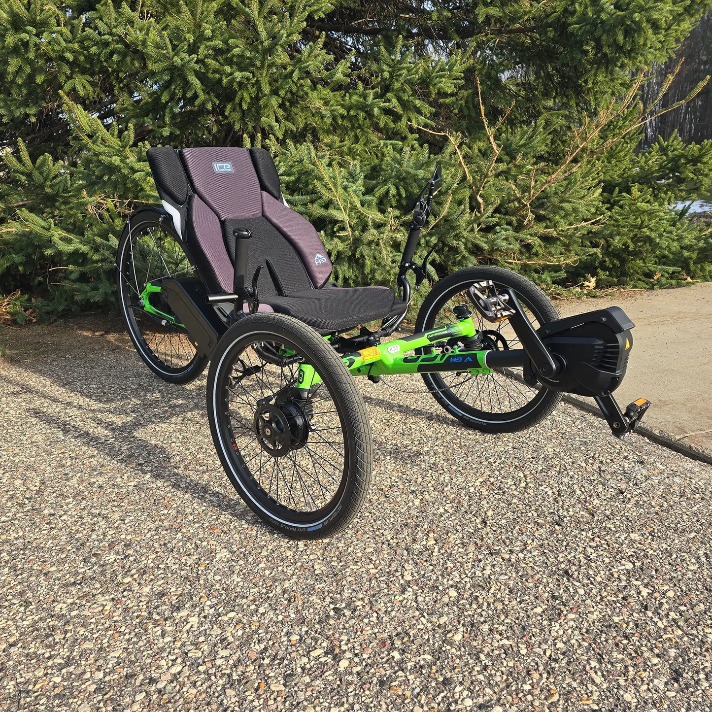 Angled view of the ICE Adventure HD 26 FS PERS Chainless Kiwi Green Recumbent Trike on a bike path near a pine tree.