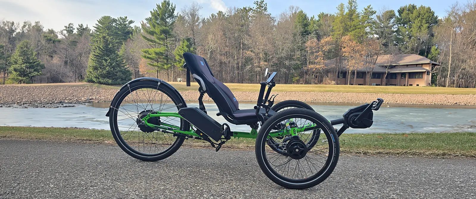 The ICE Adventure HD 26 FS PERS Chainless Kiwi Green Recumbent Trike on a bike path alongside a melting pond with trees in the background.