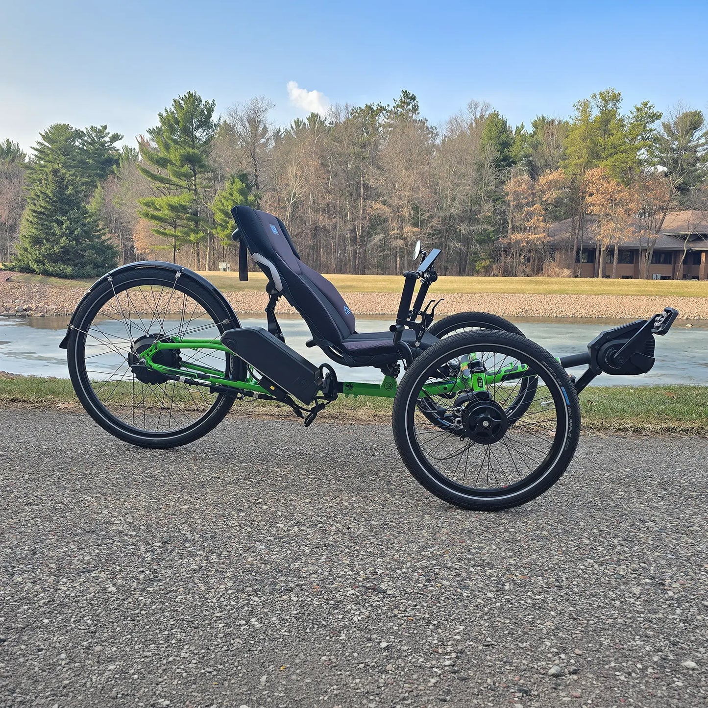 Side view of the ICE Adventure HD 26 FS PERS Chainless Kiwi Green Recumbent Trike on a bike path near a melting pond with trees in the background.